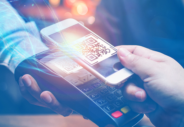 Close-up of a contactless payment. A hand holds a smartphone near a payment terminal that displays a QR code. 